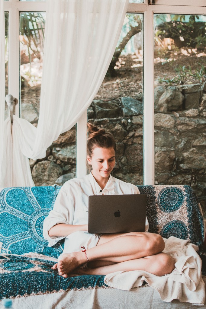 A woman working on a laptop while sitting cross-legged on a sofa beside a bright window.